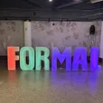 Large glow letters spelling FORMAL lit in orange, green and purple against a textured wall at a school formal