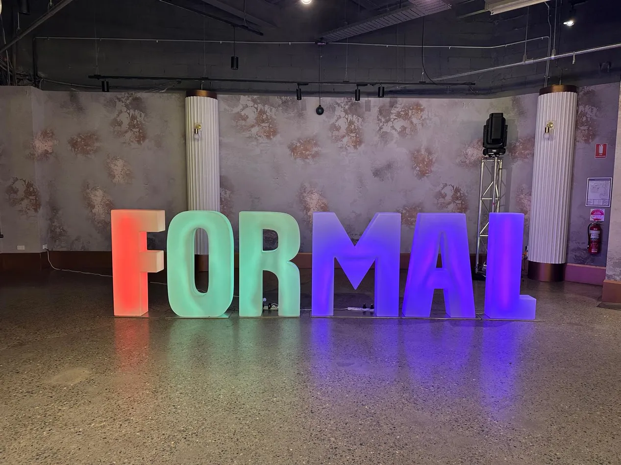 Large glow letters spelling FORMAL lit in orange, green and purple against a textured wall at a school formal