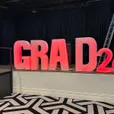 Illuminated glow letters and numbers spelling GRAD 26 lit in red across the front of a stage at a graduation event