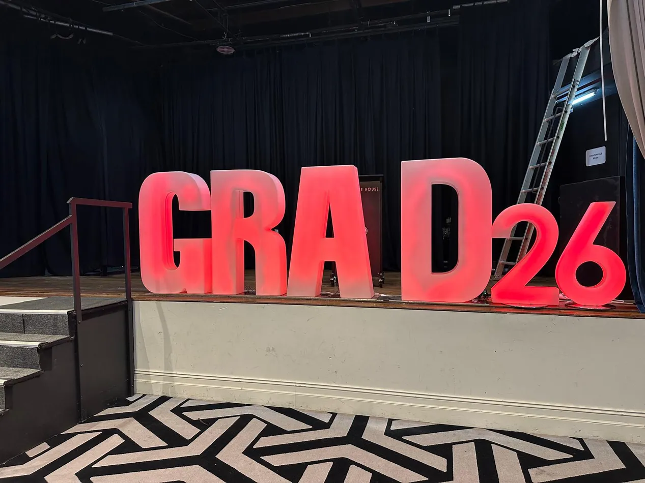 Illuminated glow letters and numbers spelling GRAD 26 lit in red across the front of a stage at a graduation event