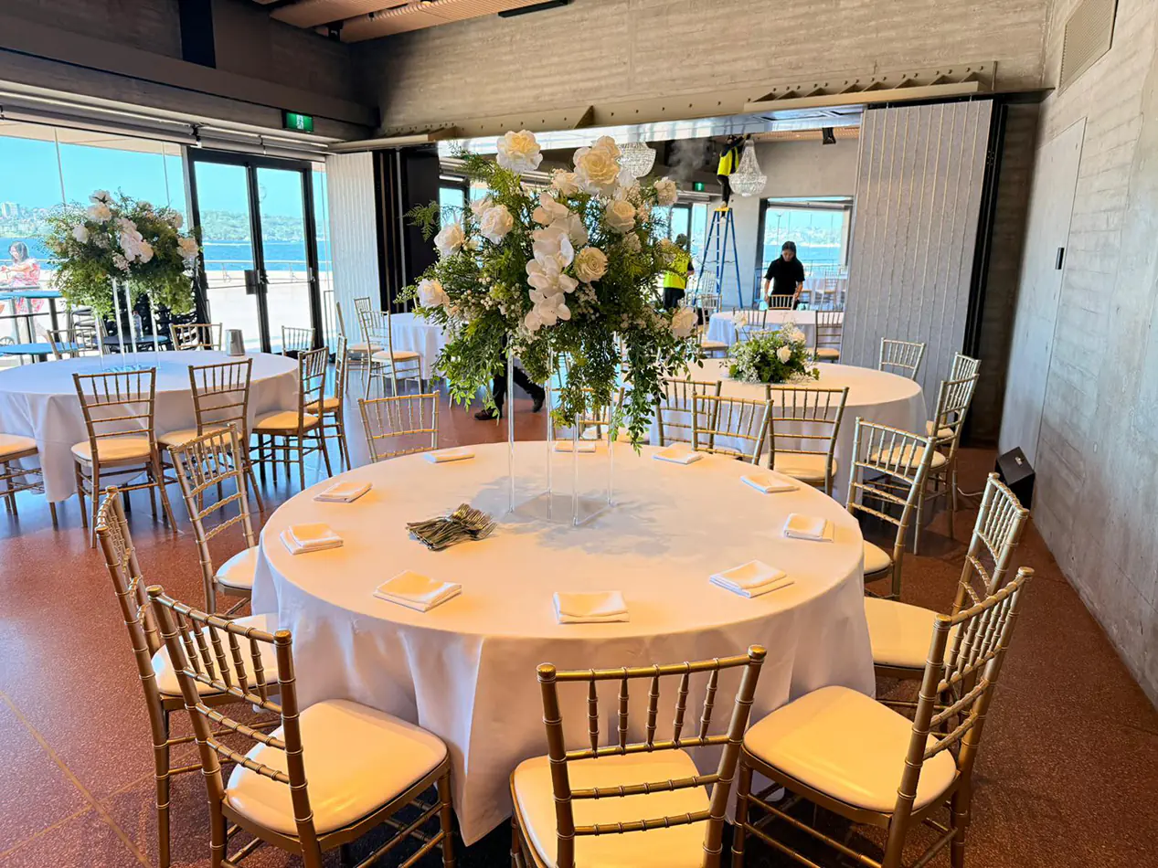 Gold Tiffany chairs arranged around a round banquet table with white linen and tall floral centrepiece in a bright event venue