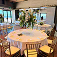 Gold Tiffany chairs arranged around a round banquet table with white linen and tall floral centrepiece in a bright event venue