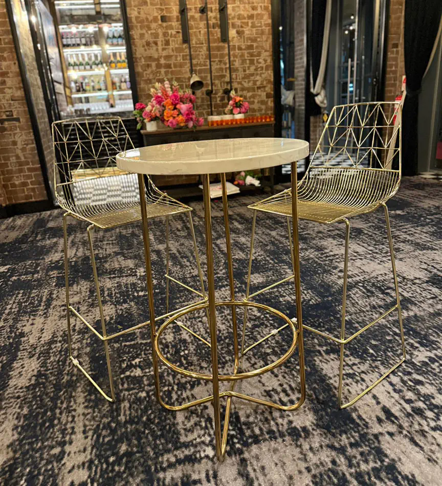 Gold wire cocktail table with marble top paired with matching gold wire stools in an indoor event space with exposed brick walls and patterned carpet
