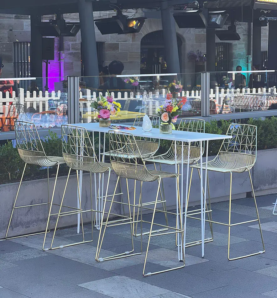 Gold wire stools arranged around a white bar table in an outdoor dining setting with floral centrepieces and glass balustrade backdrop