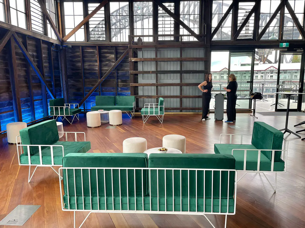 Green velvet wire lounge furniture with white ottoman stools and round coffee tables inside a timber-clad harbourfront function venue