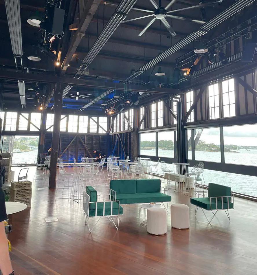 Green velvet wire lounge seating with white wire frame furniture and ottoman stools styled inside a large waterfront event venue with timber floors and harbour views