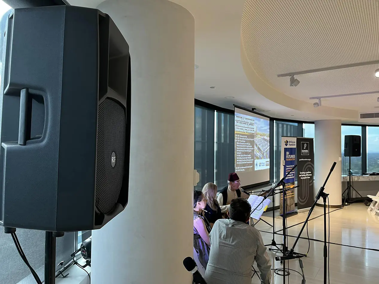 Indoor event setup with PA speaker on stand, projection screen presentation, microphones, and seated attendees in modern function room