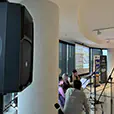 Indoor event setup with PA speaker on stand, projection screen presentation, microphones, and seated attendees in modern function room