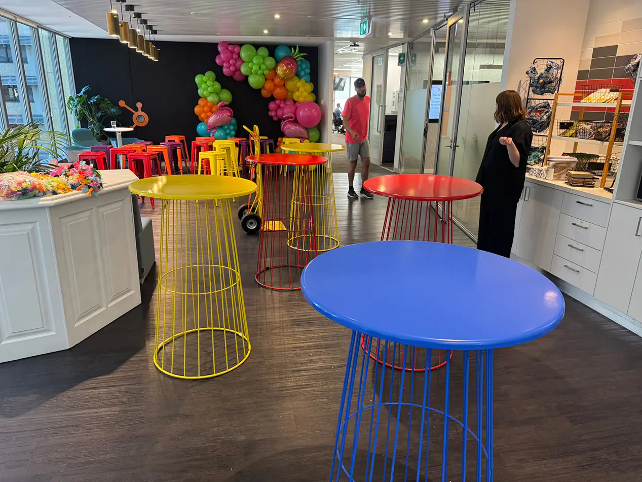 Indoor event space with colourful wire cocktail tables and stools set up for a party with balloon decorations in the background