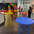 Indoor event space with colourful wire cocktail tables and stools set up for a party with balloon decorations in the background