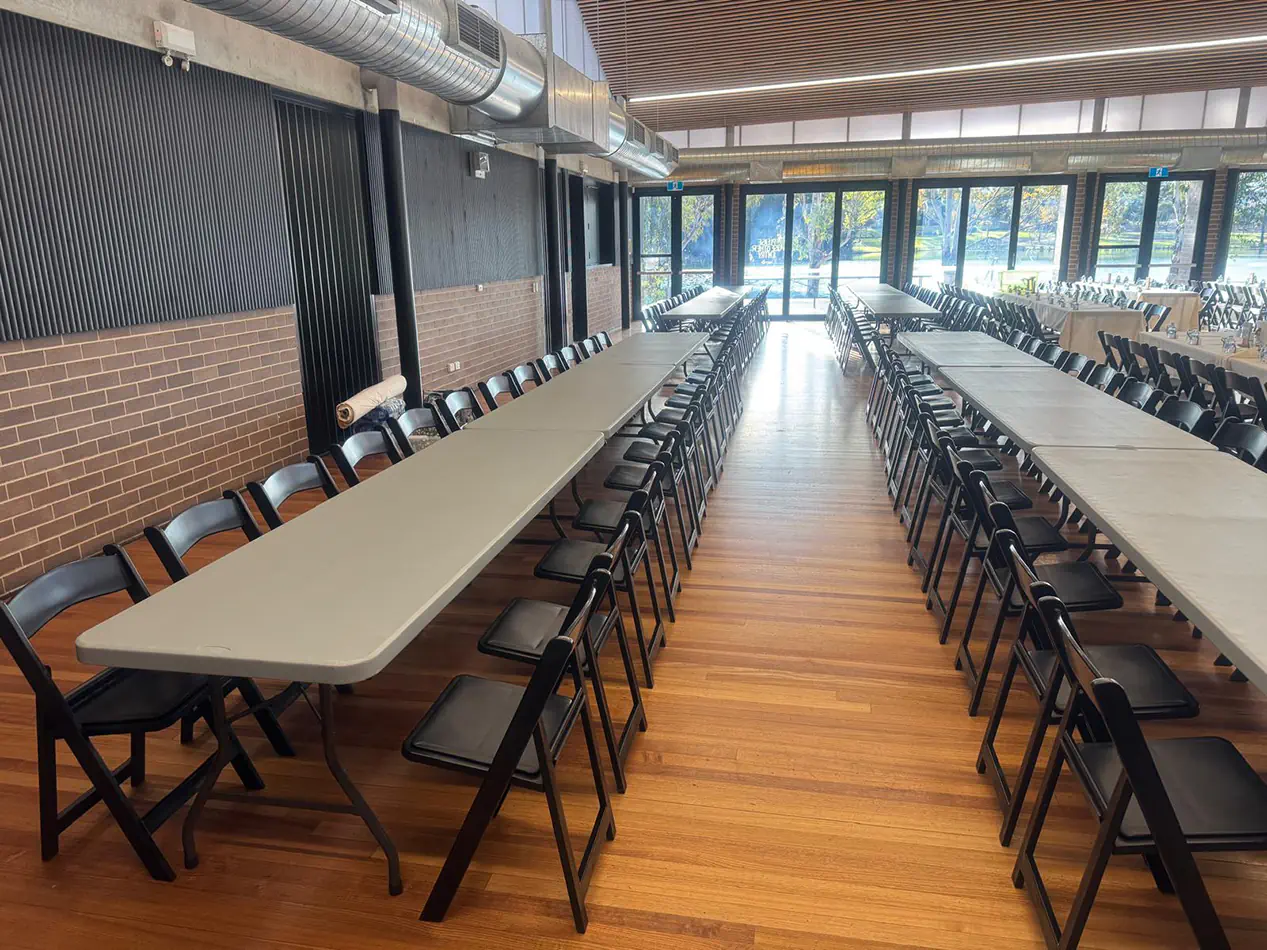 Indoor function venue with long grey trestle tables and black folding chairs arranged for a large event or presentation