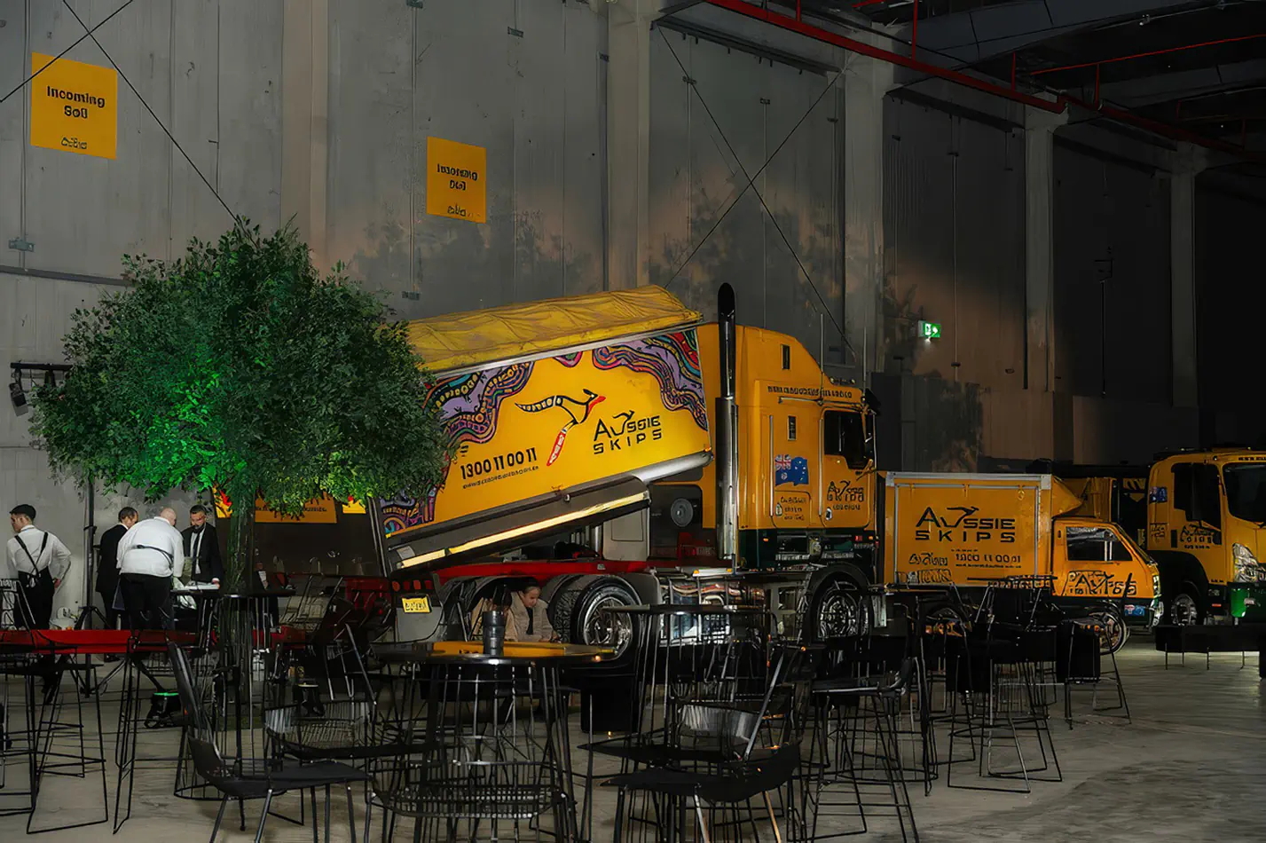 Black wire cocktail tables arranged in a large industrial venue with feature lighting, decorative tree and display trucks in the background