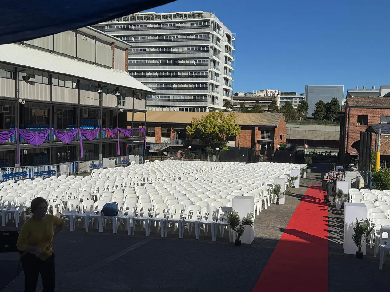 Large outdoor event setup with rows of white plastic chairs facing a red carpet aisle in a courtyard venue