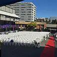 Large outdoor event setup with rows of white plastic chairs facing a red carpet aisle in a courtyard venue