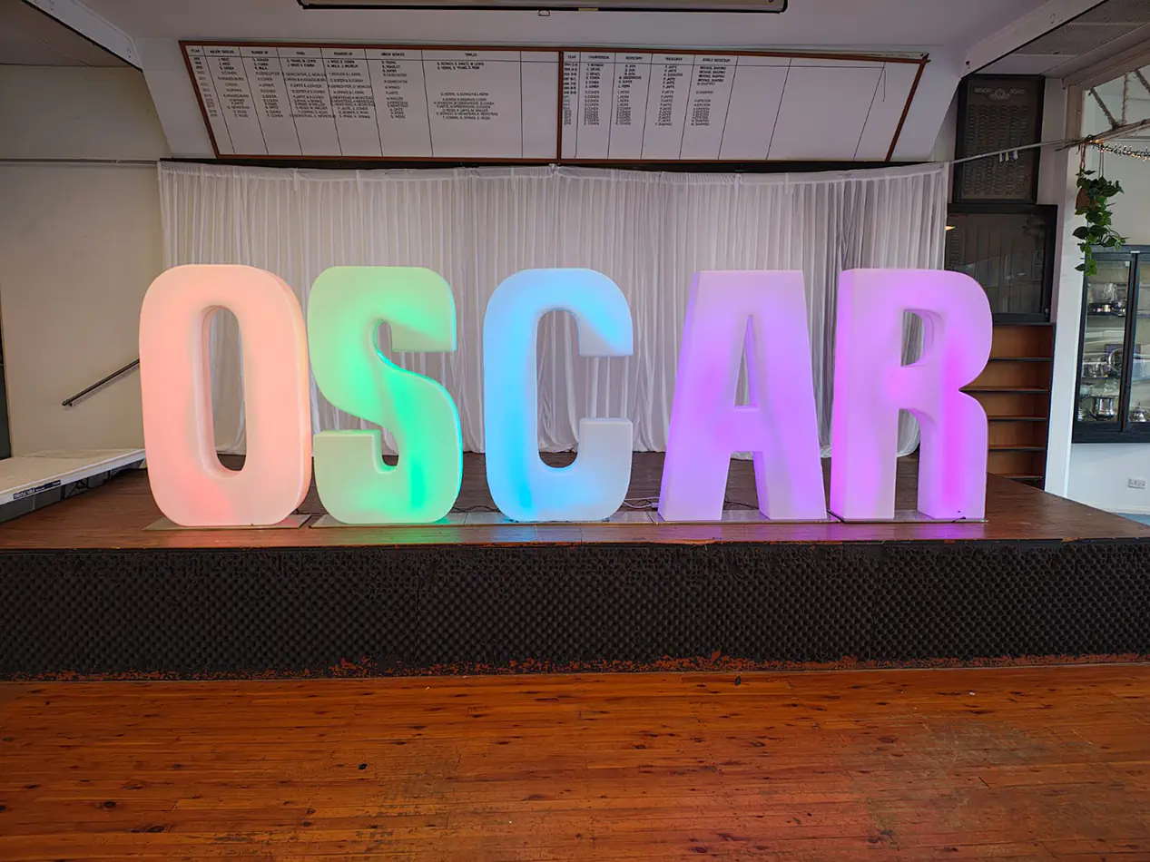 Large illuminated letter sign spelling OSCAR with soft colour lighting set on a stage with curtains and wooden flooring for an event display