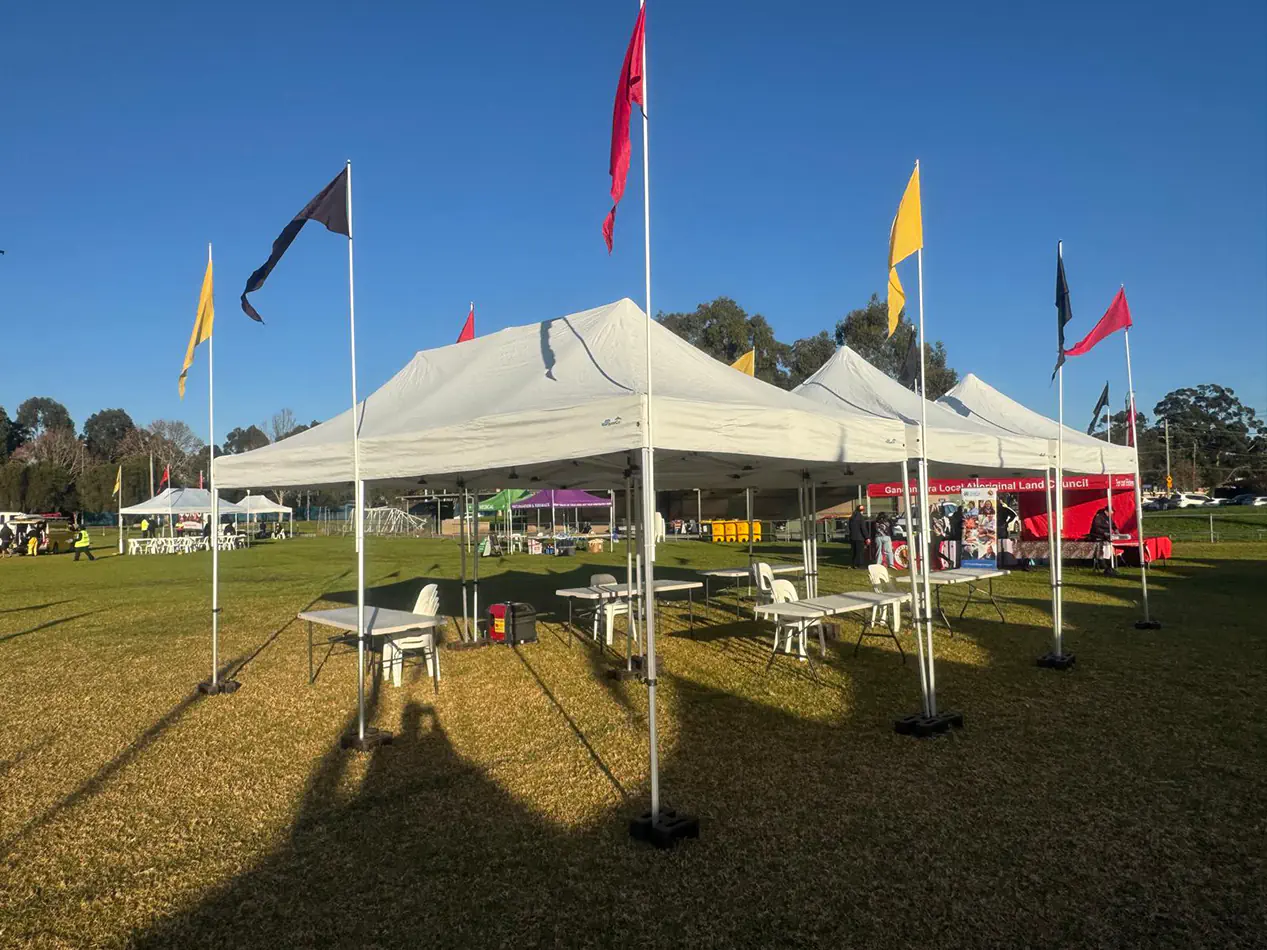 Large white pop-up marquee setup with tables, chairs and colourful flags at a community outdoor event on grass