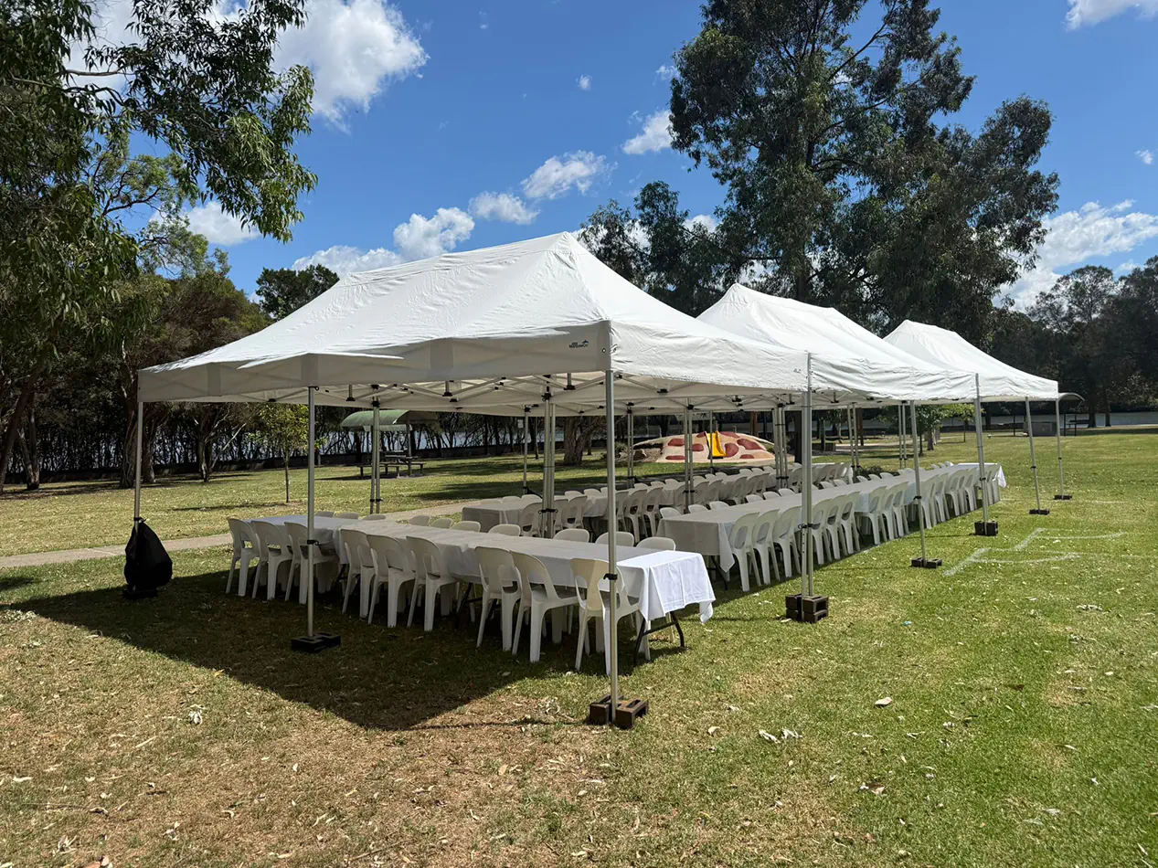 Large white pop-up marquees covering banquet tables and white chairs for an outdoor function in a park setting