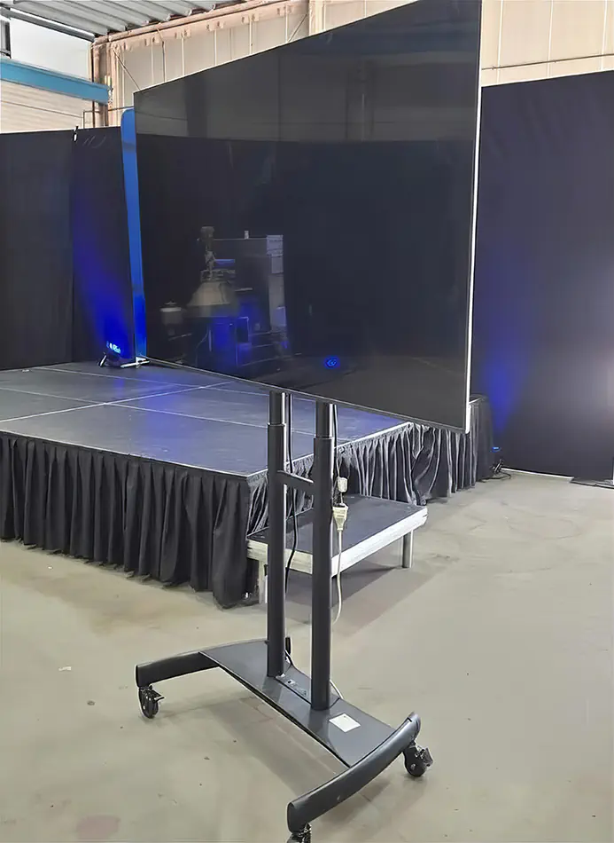 Large TV screen hire setup on a wheeled stand beside a raised event stage with black draping, lighting and warehouse-style venue backdrop