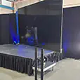 Large TV screen hire setup on a wheeled stand beside a raised event stage with black draping, lighting and warehouse-style venue backdrop