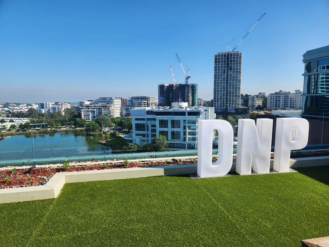 Large white illuminated letters spelling DNP positioned on a rooftop event space overlooking a city skyline with buildings and water views