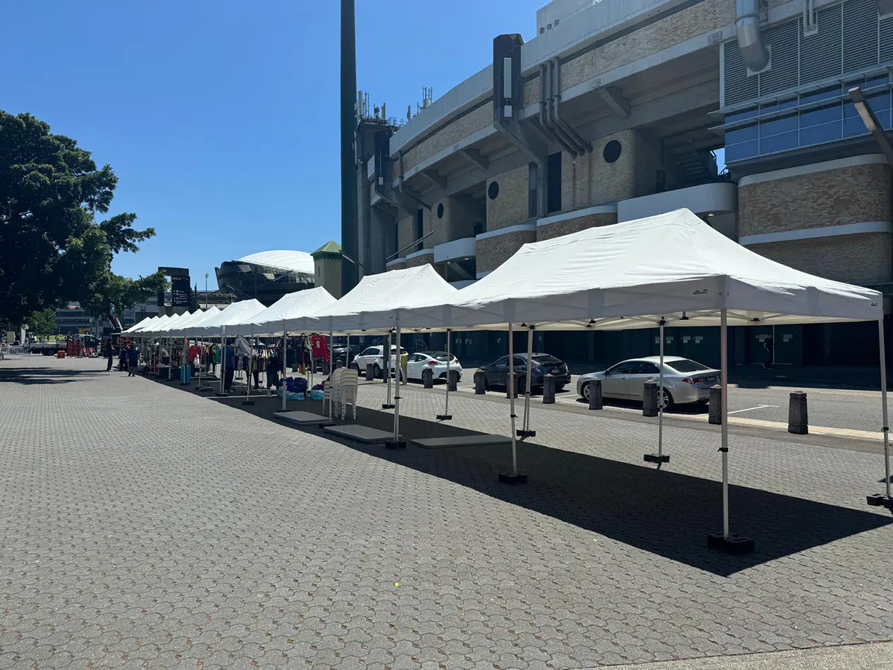 Large white pop up marquee set up on paved ground outside a stadium, with weighted legs and multiple market-style marquees in the background