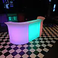 LED glow bar with curved modular sections illuminated in changing colours, set in a dark indoor event space with checkerboard flooring