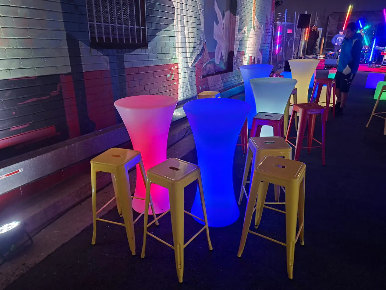 LED glow cocktail tables in vibrant colours including blue, pink, and green set along an outdoor night event space with coloured lighting and bar stools
