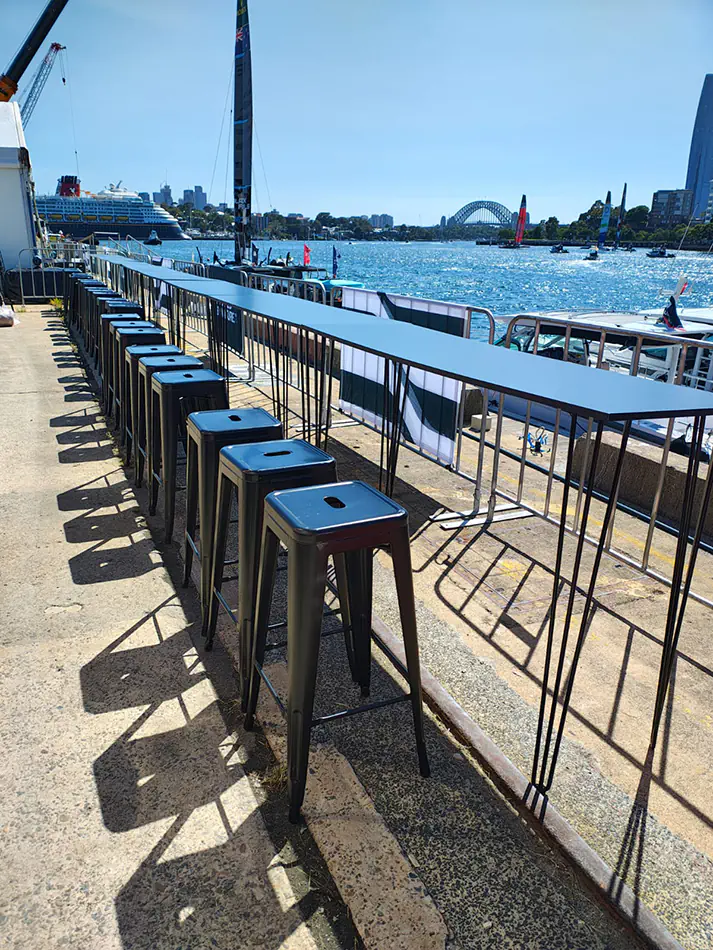 Long bar height table with black stools set up along a waterfront location overlooking Sydney Harbour with boats and bridge views in the background