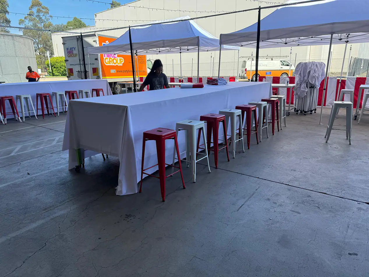 Long rectangular table covered with white linen surrounded by red and white stools under pop-up marquees in an outdoor event setup