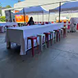 Long rectangular table covered with white linen surrounded by red and white stools under pop-up marquees in an outdoor event setup