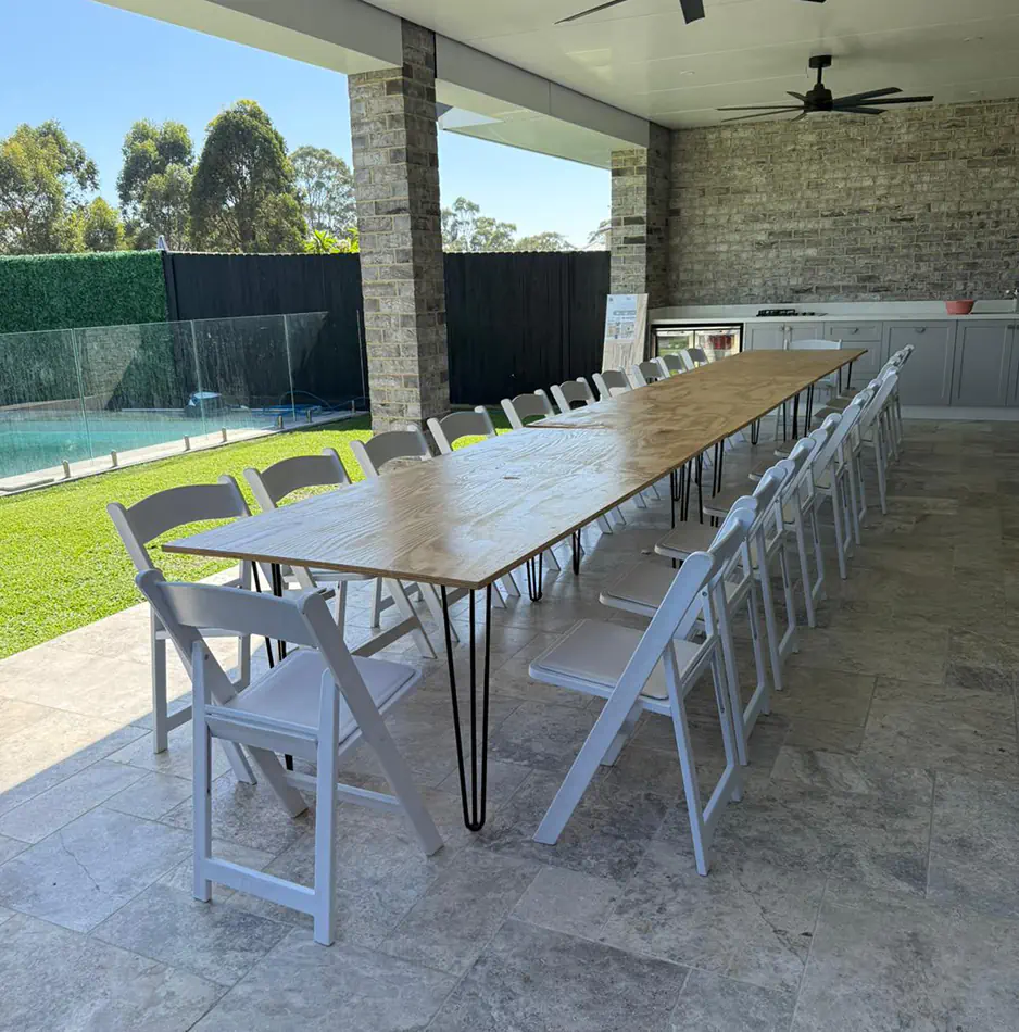 Long timber banquet table with black hairpin legs and white folding chairs on a covered outdoor entertaining patio