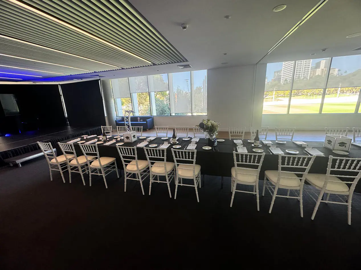 Long trestle table covered in black linen with white chairs set for a formal event inside a bright function room with large windows and stage area