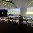 Long trestle table covered in black linen with white chairs set for a formal event inside a bright function room with large windows and stage area