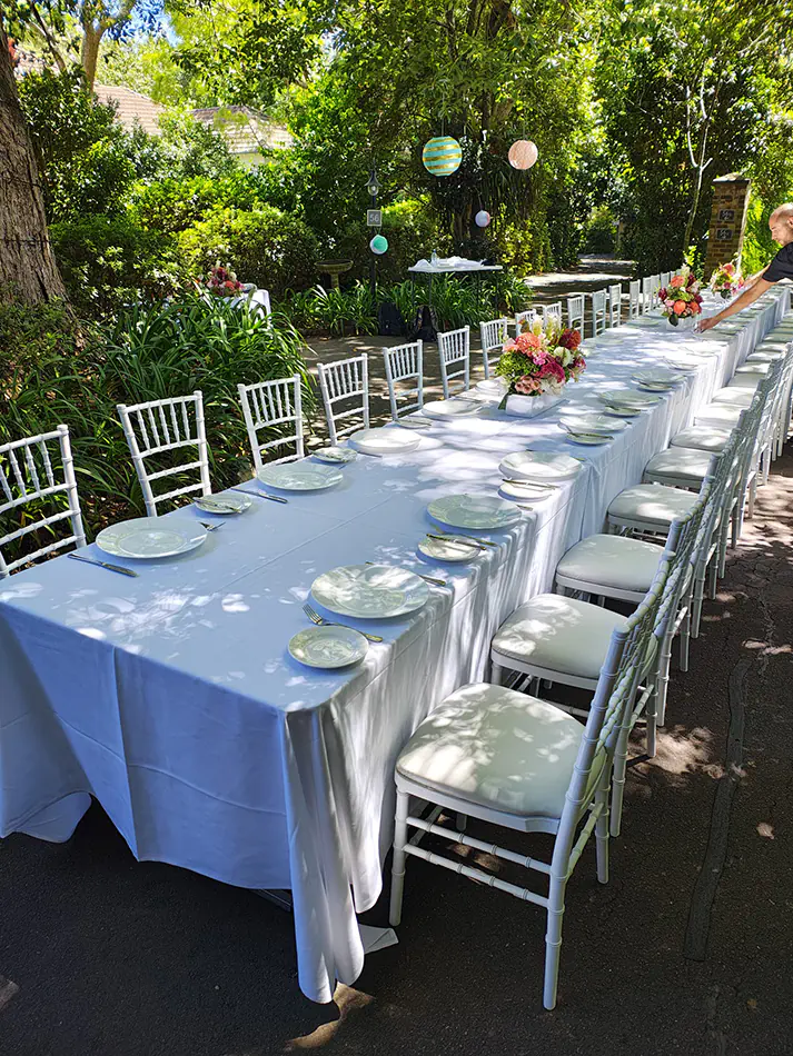 Long outdoor dining table with white linen, place settings, and white Tiffany chairs arranged in a garden setting with floral centrepieces