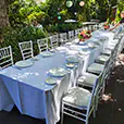 Long outdoor dining table with white linen, place settings, and white Tiffany chairs arranged in a garden setting with floral centrepieces