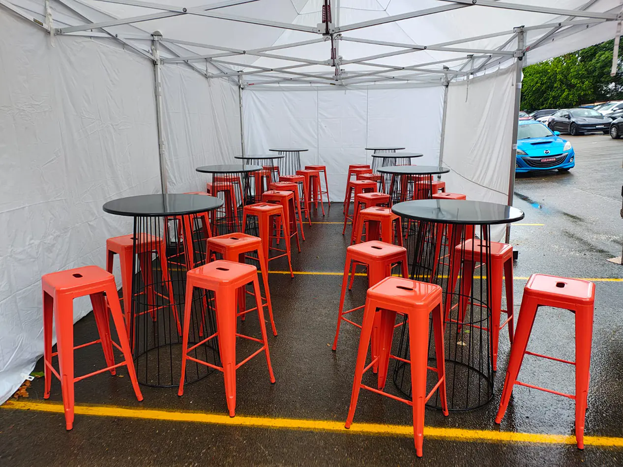 Marquee event setup with red metal bar stools and round high tables arranged under a white canopy on wet pavement