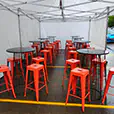 Marquee event setup with red metal bar stools and round high tables arranged under a white canopy on wet pavement