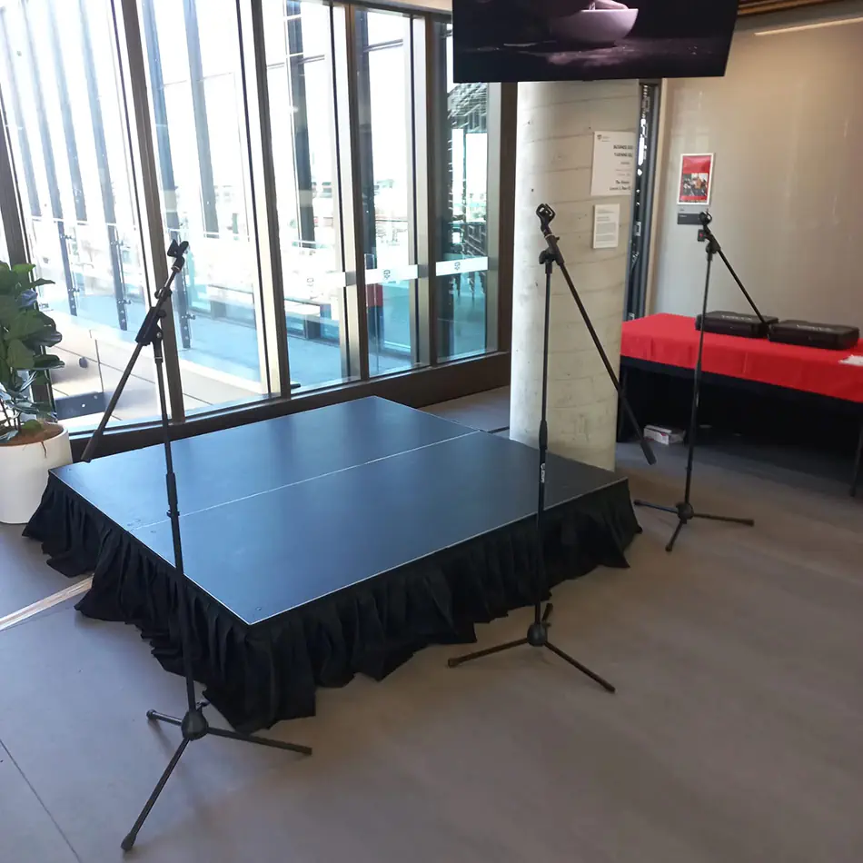 Small indoor stage setup with black stage platform and two microphone stands positioned for presentations or performances inside a modern event venue with large windows