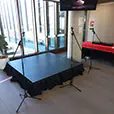 Small indoor stage setup with black stage platform and two microphone stands positioned for presentations or performances inside a modern event venue with large windows