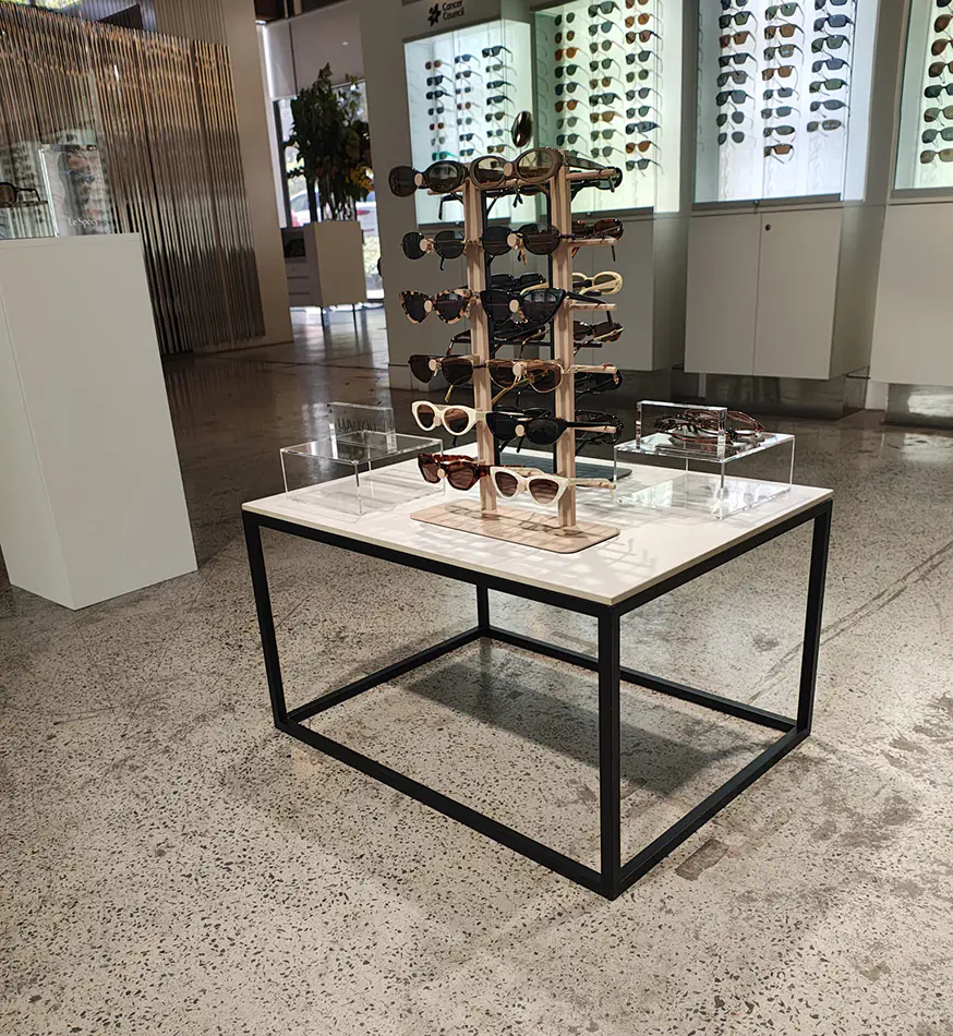 Modern display table with black frame and white top showcasing sunglasses in a retail showroom with illuminated wall displays