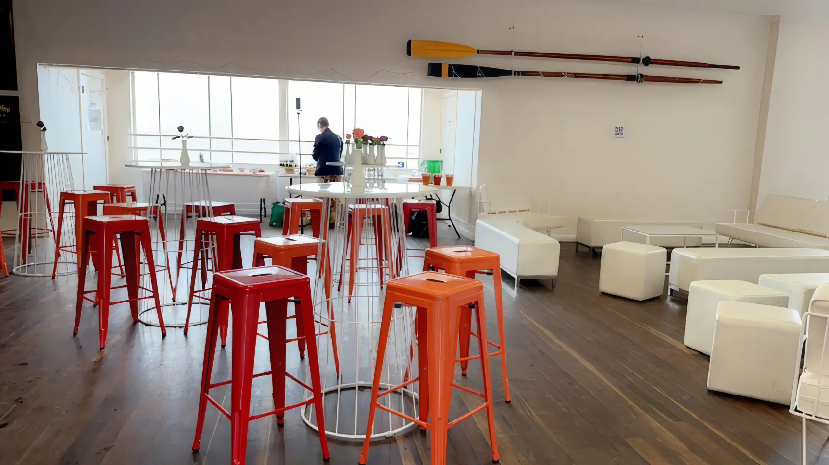 Orange tolix stools arranged around white cocktail tables in a modern indoor event space with timber flooring and lounge seating area