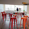 Orange tolix stools arranged around white cocktail tables in a modern indoor event space with timber flooring and lounge seating area