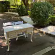 Ornate vintage style table with classic drawer detailing, placed with two folding white chairs on a shaded garden pathway lined with shrubs and white gravel.