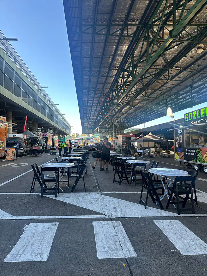 Outdoor cafe tables and folding chairs arranged along a street-style market setup with food trucks and covered structure overhead
