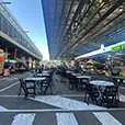 Outdoor cafe tables and folding chairs arranged along a street-style market setup with food trucks and covered structure overhead
