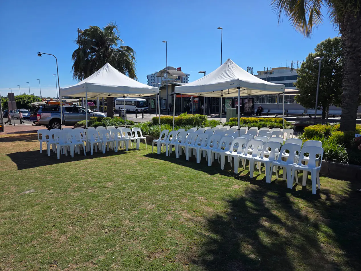 Outdoor ceremony setup with white pop-up marquees and rows of white chairs arranged on grass near a transport precinct