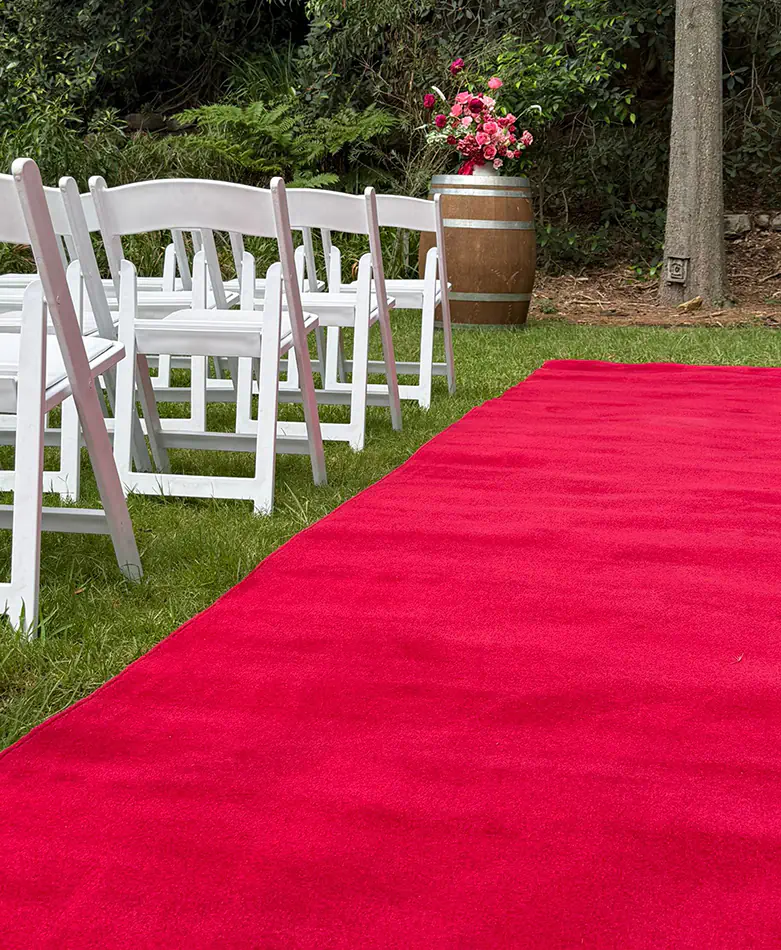 Outdoor ceremony setup with red carpet aisle, garden surroundings, floral arrangement, and guest seating for a formal event