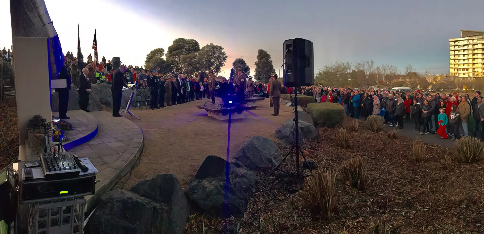 Outdoor ceremony with large crowd, PA speaker on stand, audio mixer setup, and microphone podium during public event