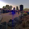 Outdoor ceremony with large crowd, PA speaker on stand, audio mixer setup, and microphone podium during public event