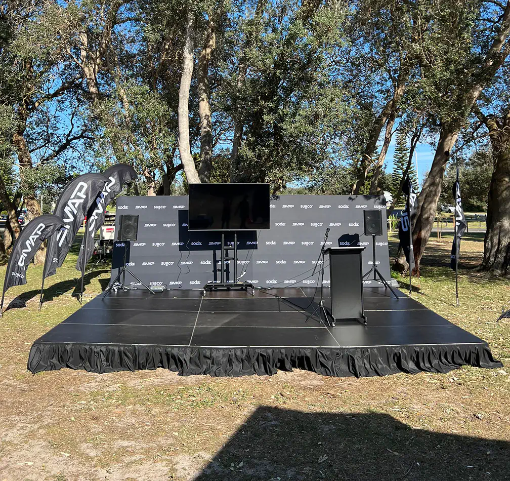 Outdoor event stage setup with large TV screen, speakers, microphone stand, and branded backdrop surrounded by trees in a park setting